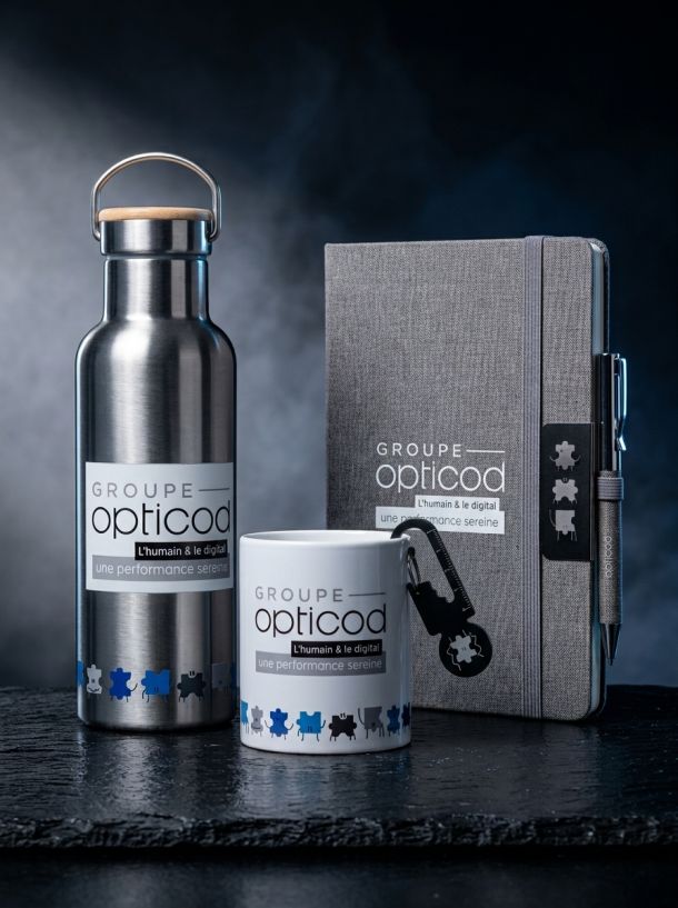 Objets publicitaires personnalisés Groupe Opticod : gourde inox, mug, carnet de notes gris et stylo sur fond sombre.